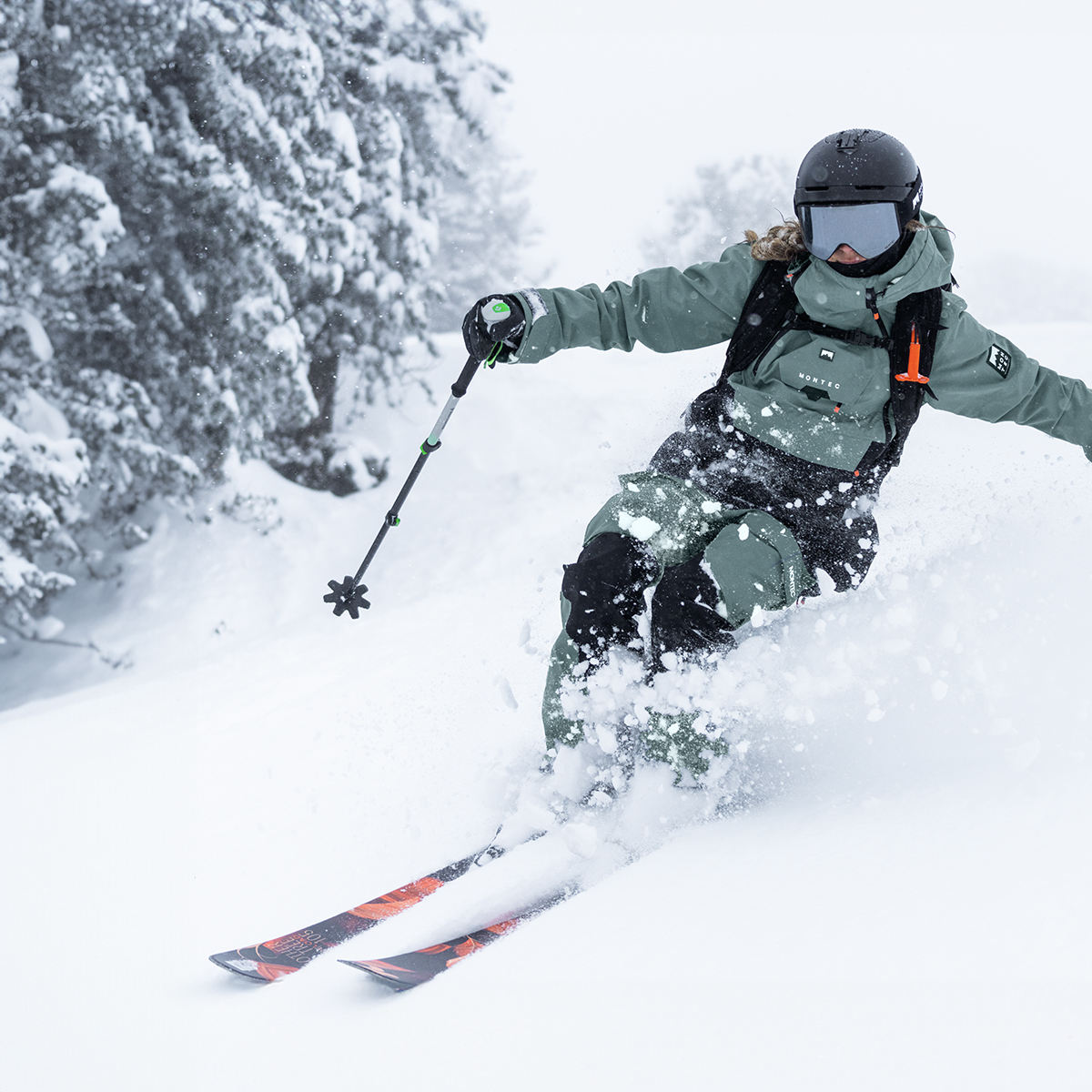 A skier, dressed in a green winter montec outfit with a helmet and goggles, carves through fresh powder on a snowy slope. Snow sprays around them as they navigate the terrain with ski poles, with snow-covered trees visible in the background under a soft, overcast sky.